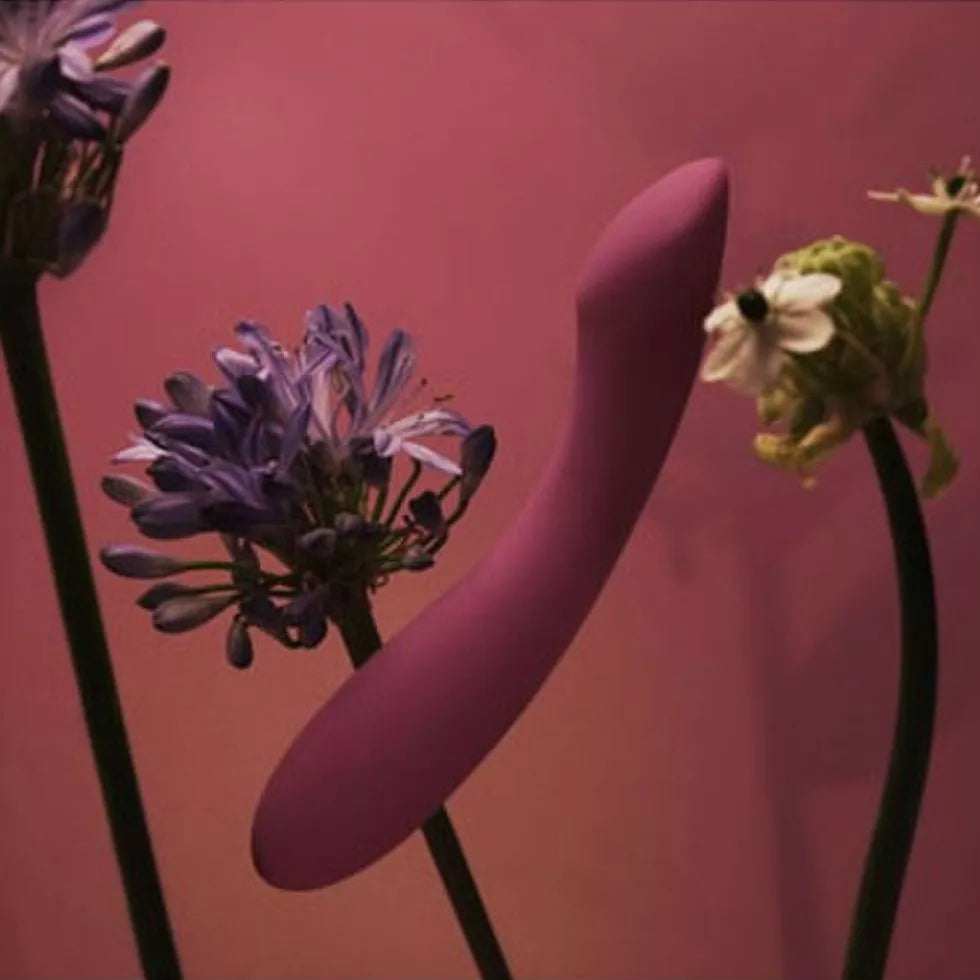The Svakom Amy 2 – Strong Flexible G-Spot Vibrator, a purple body-safe silicone toy by Svakom, is displayed among purple and green flowers on a pink background.