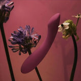The Svakom Amy 2 – Strong Flexible G-Spot Vibrator, a purple body-safe silicone toy by Svakom, is displayed among purple and green flowers on a pink background.