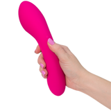 A hand with pink-painted nails holds The Swan Wand, a pink curved vibrator by the brand Swan, featuring a smooth texture. The device has a bulbous end and tapers slightly towards the middle. The background is plain white.