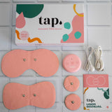 Contents of the Tap. 2 Wearable TENS Machine set from Tap Health displayed on a tiled surface include a blush pink wearable TENS device, electrode pads, charging cables, a user manual, and a decorative box with colorful abstract patterns for personalized pain relief.