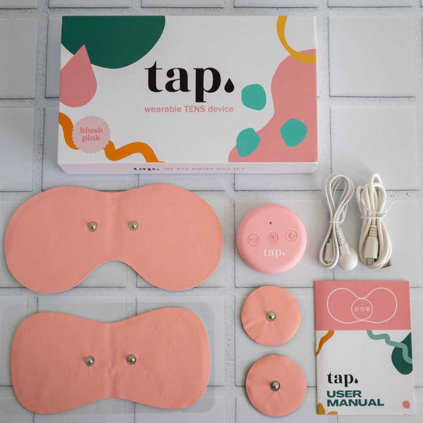 Contents of the Tap. 2 Wearable TENS Machine set from Tap Health displayed on a tiled surface include a blush pink wearable TENS device, electrode pads, charging cables, a user manual, and a decorative box with colorful abstract patterns for personalized pain relief.