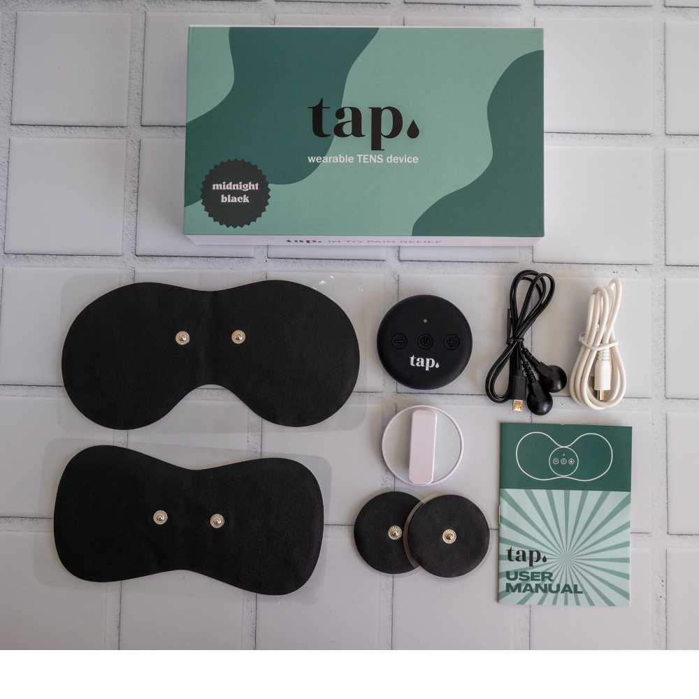 A packaging box with a green and white design, labeled "Tap. 2 Wearable TENS Machine" from Tap Health, is surrounded by components such as adhesive pads, a round control unit, charging cables, and a user manual. This portable pain relief solution is neatly arranged on a tiled surface.