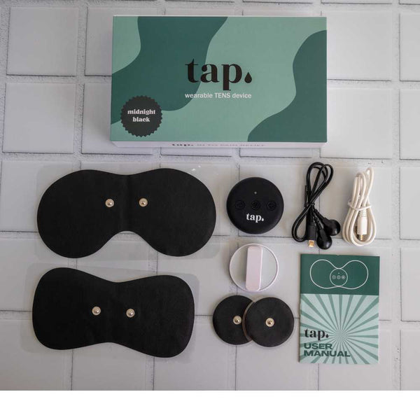 A packaging box with a green and white design, labeled "Tap. 2 Wearable TENS Machine" from Tap Health, is surrounded by components such as adhesive pads, a round control unit, charging cables, and a user manual. This portable pain relief solution is neatly arranged on a tiled surface.