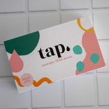 The blush pink box of the Tap. 2 Wearable TENS Machine from Tap Health features a chic design with abstract shapes in green, pink, and orange. This portable pain relief solution is beautifully packaged on a tiled surface, prepared to deliver personalized comfort wherever you need it.