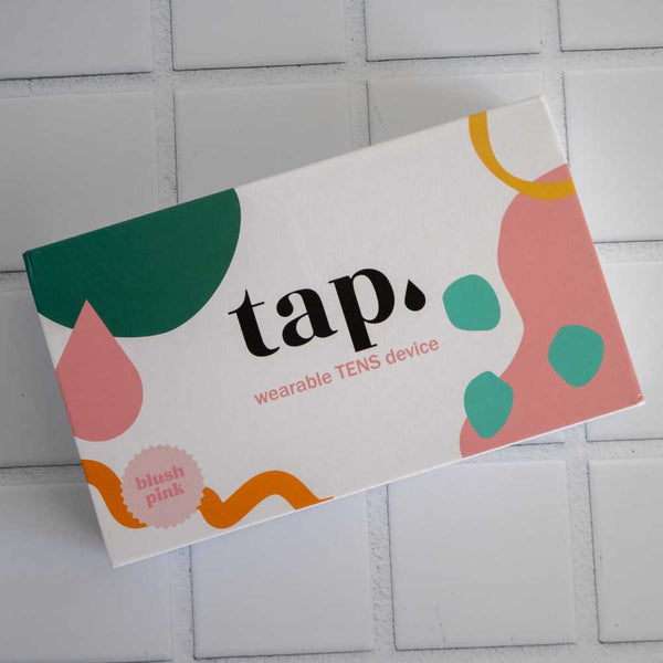 The blush pink box of the Tap. 2 Wearable TENS Machine from Tap Health features a chic design with abstract shapes in green, pink, and orange. This portable pain relief solution is beautifully packaged on a tiled surface, prepared to deliver personalized comfort wherever you need it.