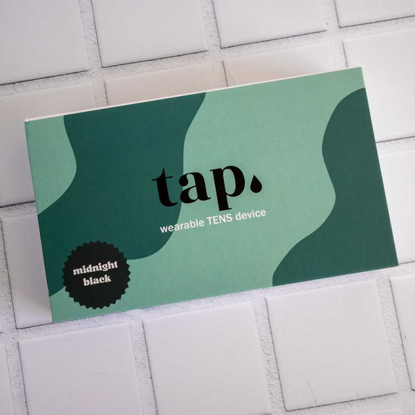 A rectangular box on a tiled surface labeled "Tap. 2 Wearable TENS Machine," showcasing a color scheme of various shades of green and adorned with a black sticker that reads "midnight black," promises personalized pain relief with its sleek, portable design by Tap Health.