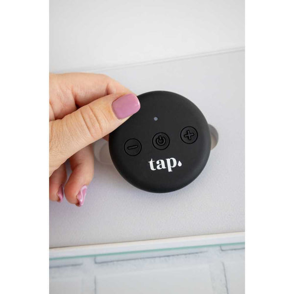 A hand is holding a Tap. 2 Wearable TENS Machine, a round, black device from Tap Health with buttons labeled "tap," resembling a wearable pain relief tool. One button features a power symbol, while others are marked with plus and minus signs. This portable device sits on a light surface that includes a cut-out section.
