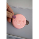 A person with a purple-painted fingernail is holding a round, pink remote for the Tap. 2 Wearable TENS Machine by Tap Health, featuring three buttons: minus, power, and plus, with the word 
