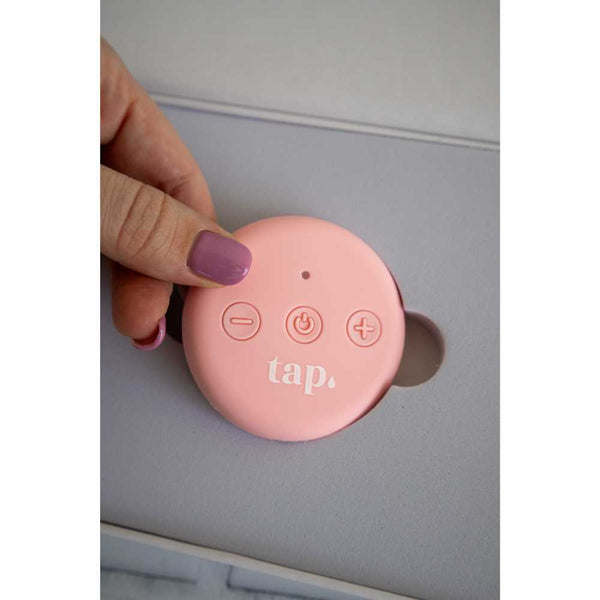 A person with a purple-painted fingernail is holding a round, pink remote for the Tap. 2 Wearable TENS Machine by Tap Health, featuring three buttons: minus, power, and plus, with the word "tap" printed on it. This device for personalized pain relief is placed in a gray holder.