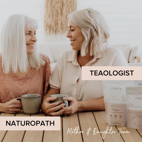 Two women sit at a wooden table, smiling at each other. The woman on the left, labeled 'Naturopath,' has long white hair and a pink top. The woman on the right, labeled 'Tealogist,' has short blonde hair and a beige top. Bags of a tea product are on the table. Text below reads "Mother & Daughter Team.