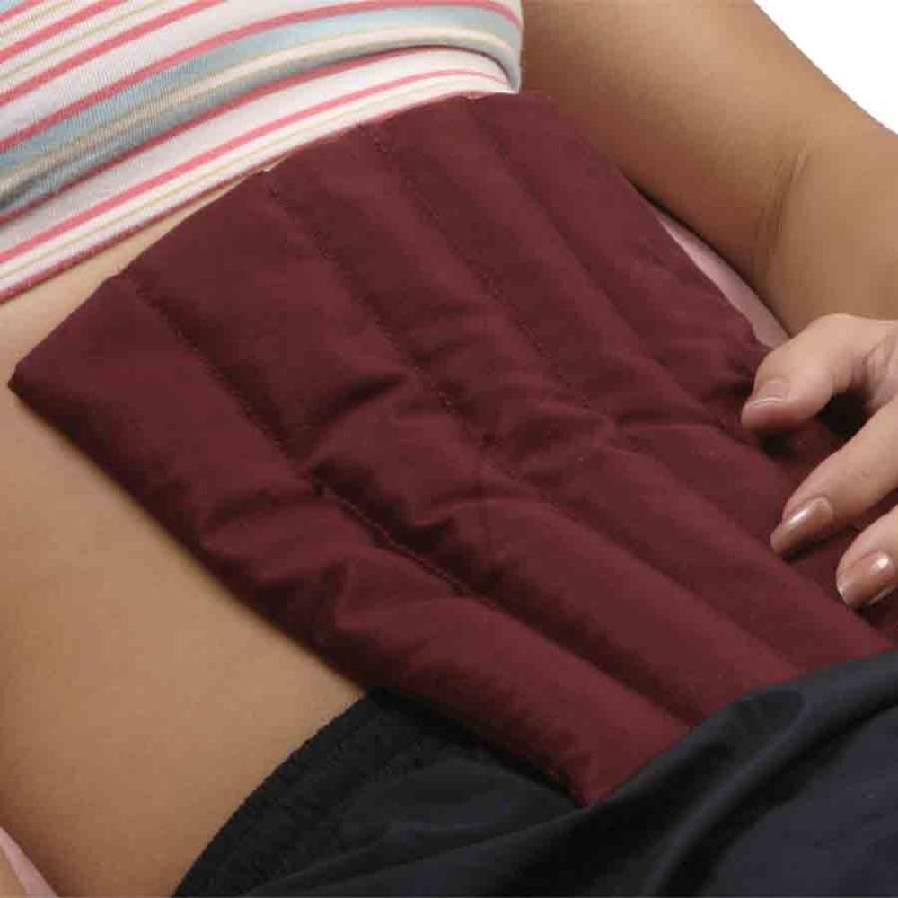 A person is lying down with a Therapack's burgundy *Therapacks Hot Pack - Back & Stomach* placed on their lower abdomen, seeking pain relief. They are wearing a striped tank top and black pants. One hand rests on their stomach near the heat pack.