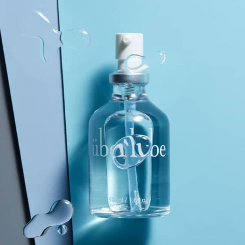 Against a blue backdrop, a clear glass spray bottle glistens with tiny water droplets. Embellished with "Uberlube," this bottle contains Uberlube, a premium silicone lubricant designed to enhance sexual wellness.
