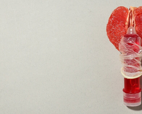 A red grapefruit slice shaped like a heart sits behind a small vial of red liquid wrapped in a condom, against a plain gray background.
