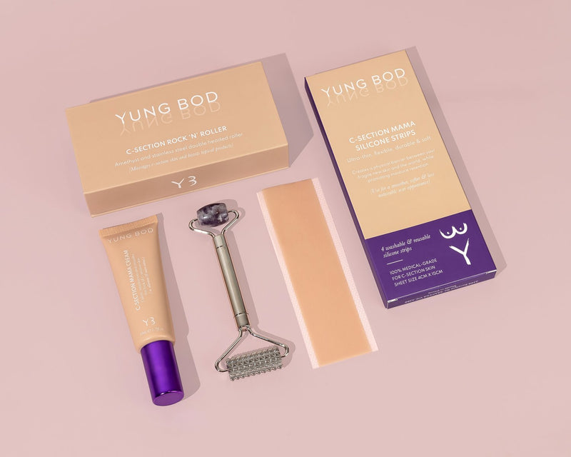 A set of skincare products on a pink surface includes a box labeled "C-Section Rock 'N' Roller," a roller tool, a tube of cream, silicone strips, and another box labeled "C-Section Mama Silicone Strips" by Yung Bod.